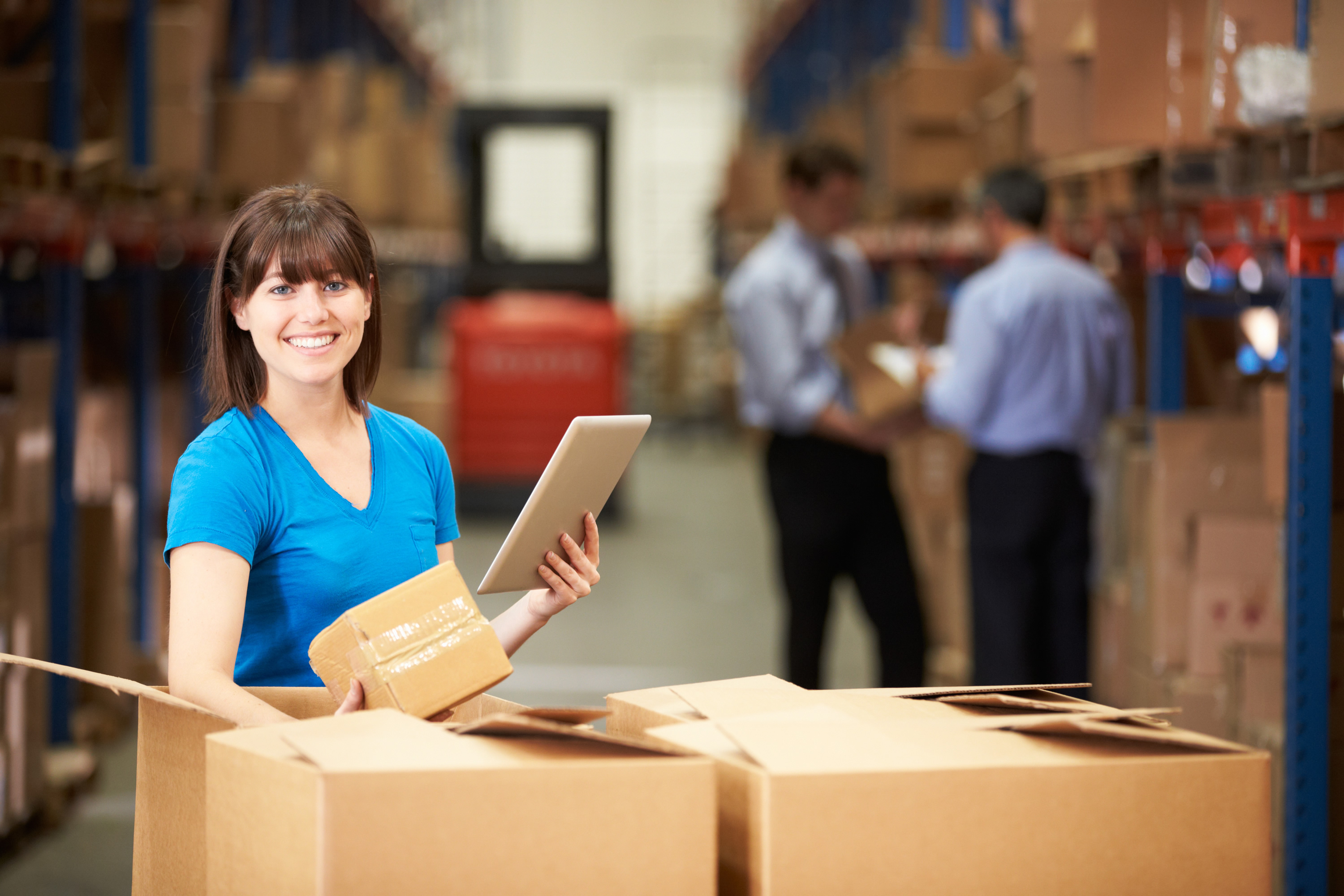 Woman in a blue shirt holding a tablet and a cardboard box in a warehouse, illustrating inventory management and operational efficiency in the printing industry.
