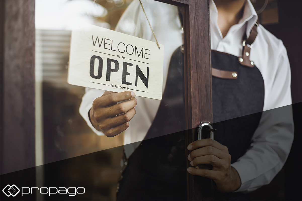 Hand holding a wooden "Welcome We Are Open Please Come In" sign, symbolizing business reopening and customer reassurance in a post-COVID context, with Propago branding visible.