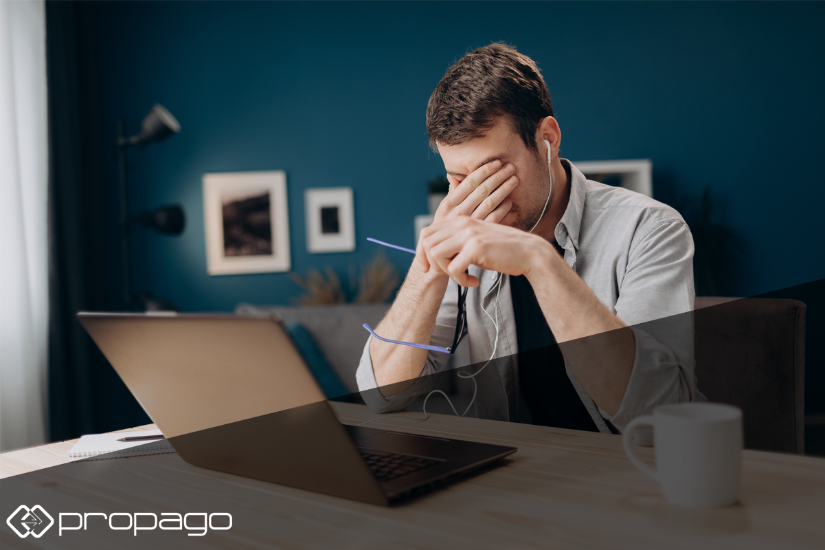 Man showing signs of digital fatigue while working on a laptop, with headphones on and hand covering face, in a home office setting.