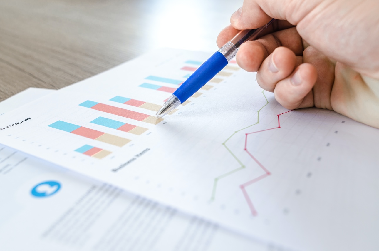 Hand holding a pen pointing at a printed graph with colorful bar charts and a line graph, illustrating data analysis relevant to B2B web portal features in the printing industry.