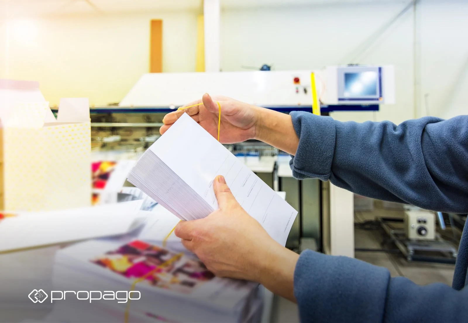 Hands holding a stack of printed materials with a yellow rubber band, in a print production environment, illustrating personalized print solutions for marketing by Propago.