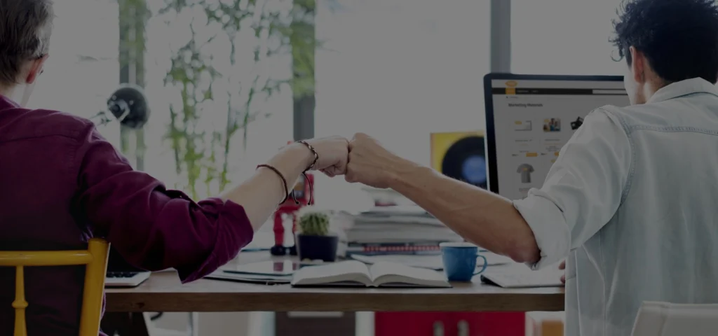 Two colleagues fist bumping at a desk with a laptop and marketing materials, symbolizing collaboration in marketing asset management.
